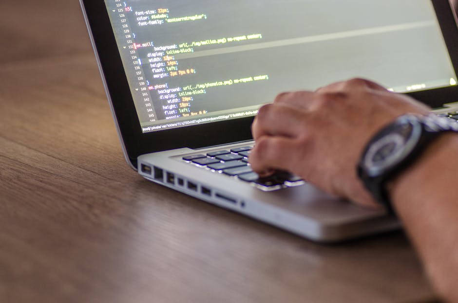A close-up of a person's hand using a laptop on a wooden desk, displaying lines of code on the screen. The individual is wearing a wristwatch and appears to be engaged in programming or web developmen